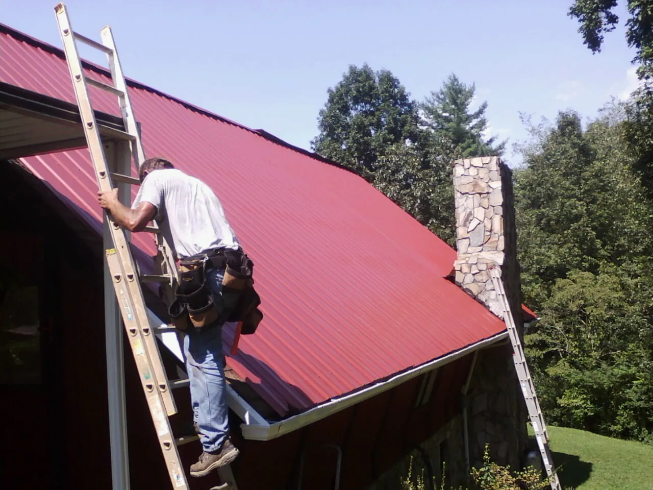 Roofer on a ladder beside a red metal roof and stone chimney — Soffit & Fascia Repair by Roofing Brothers in Mapleton